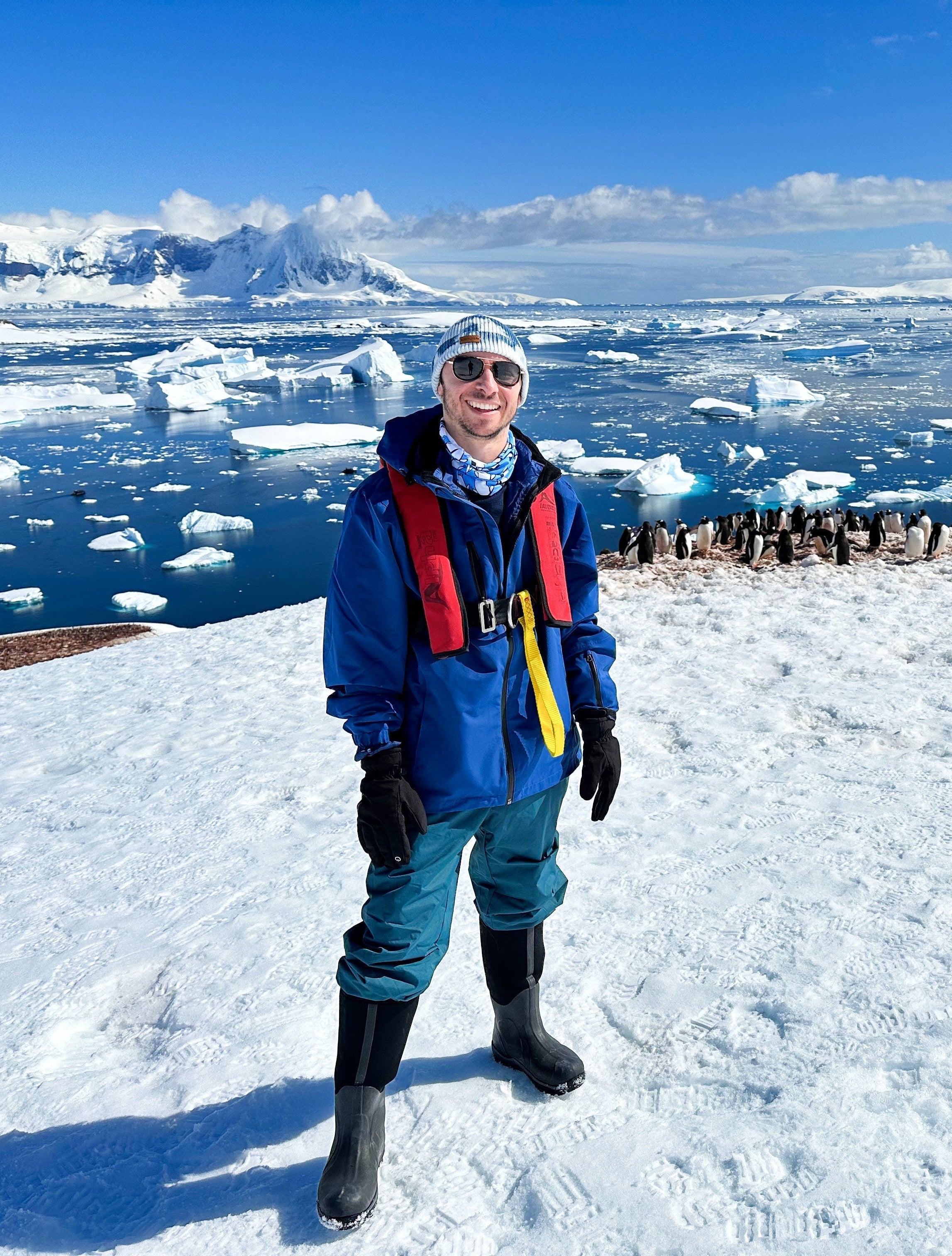 Co-founder Dani in a blue jacket and life vest on the snow, with icebergs and a penguin colony behind