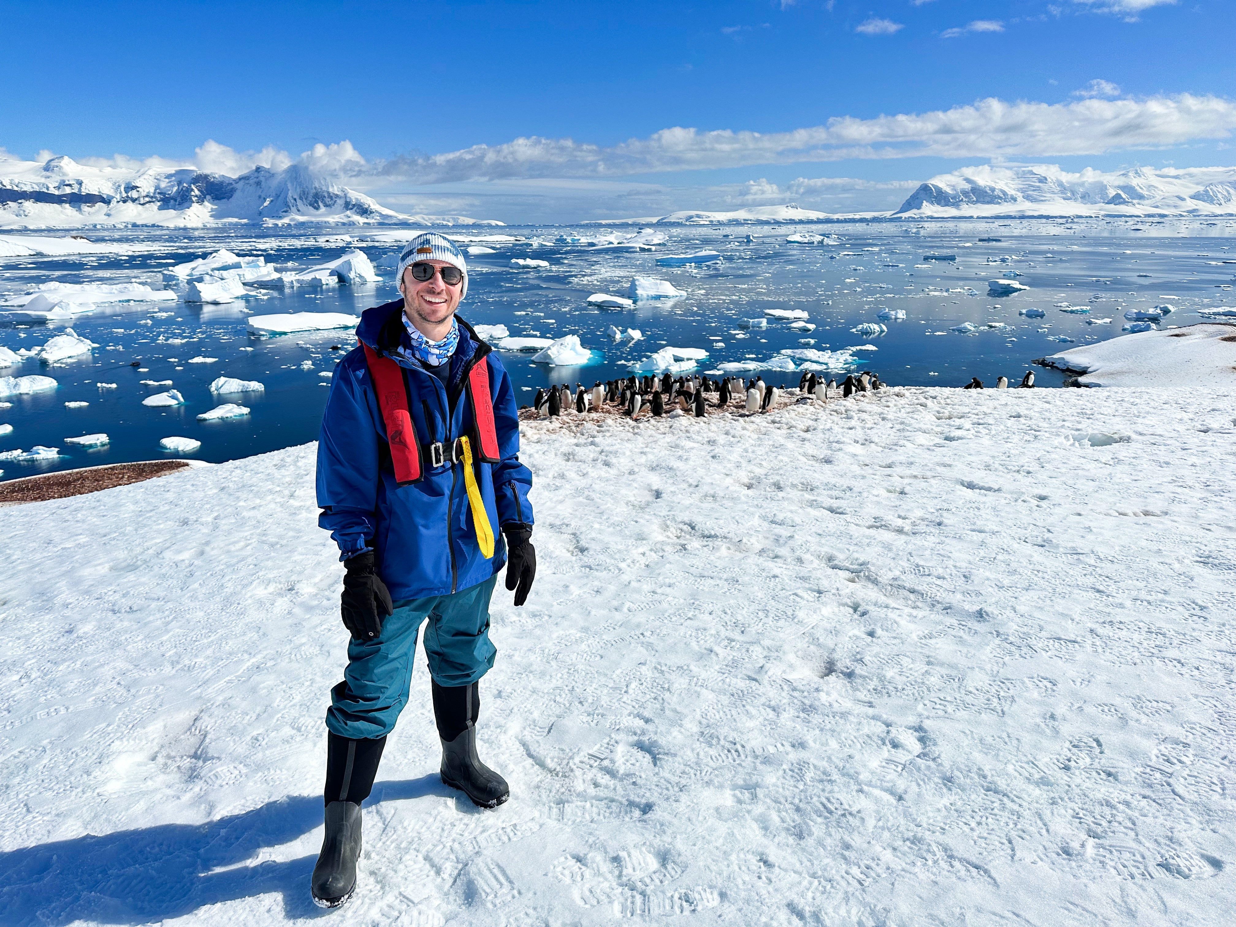 Co-founder Dani in a blue jacket and life vest on the snow, with icebergs and a penguin colony behind